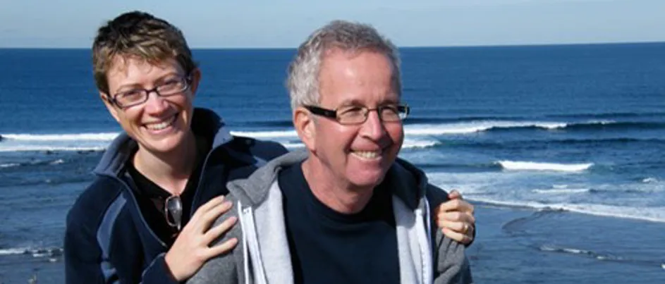 Caroline and Avner sitting together by the ocean, smiling with their arms around each other.