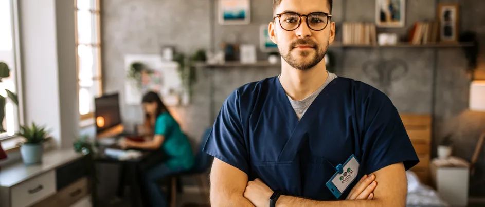 A male healthcare professional in navy blue scrubs stands confidently with arms crossed in a cozy, well-lit room. Behind him, another medical worker is seated at a desk working on a computer. The space has homey decor, including bookshelves, framed art, and natural light streaming through large windows.