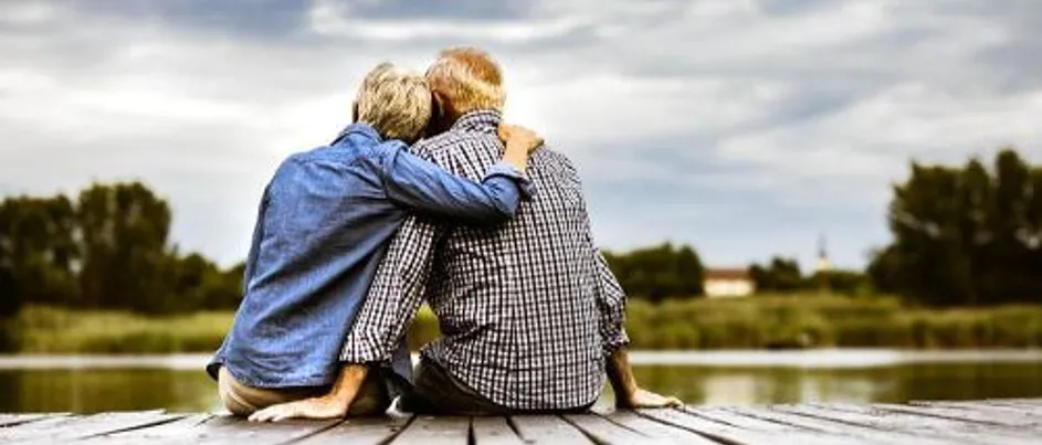 A man and a woman sitting on the side of a wooden board walk, embracing one another.