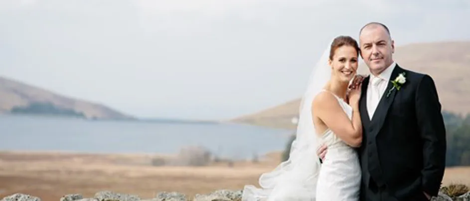George and his wife on their wedding day. They stand holding one another in front of a picturesque lake in the background.
