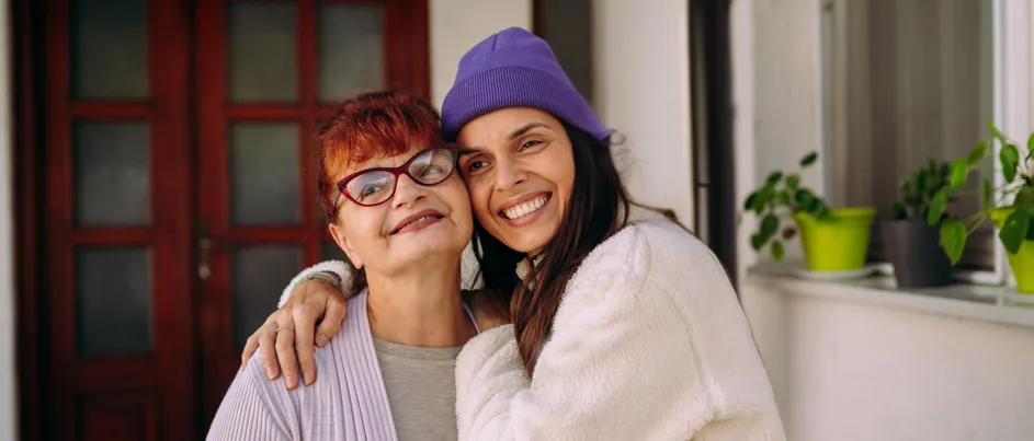 Young woman in a purple beanie hugging an older woman with red glasses, both smiling warmly in front of a house