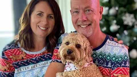 Mark, a man living with pancreatic cancer, sits smiling beside his partner in front of a Christmas tree, both wearing matching festive pyjamas. Mark is holding a fluffy tan-coloured dog.
