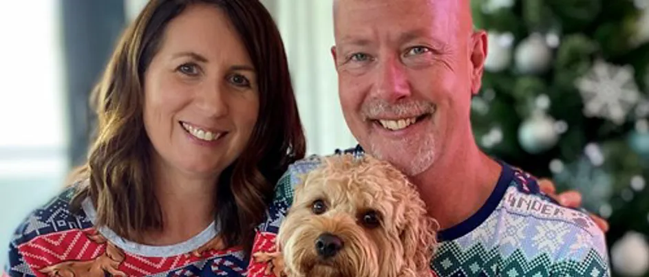 Mark, a man living with pancreatic cancer, sits smiling beside his partner in front of a Christmas tree, both wearing matching festive pyjamas. Mark is holding a fluffy tan-coloured dog. The image reflects warmth, joy, and the support of loved ones during the holiday season.